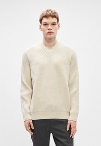 Κρεμ πλεκτή ζακέτα με V λαιμόκοψη, ριγωτή υφή και μακριά μανίκια. Διαθέτει στενή περιφέρεια. Φοριέται πάνω από σκούρες γκρι παν trousers.