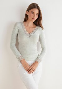 Světle šedý dlouhý rukáv top s výstřihem do V a krajkovým lemem, strukturovaná tkanina a krajkové detaily na rukávech. V kombinaci s bílými kalhotami.