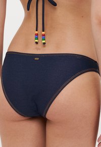 Sötétkék bikini alsó narancssárga varrással, sima textúrával és díszes, több színű gyöngyökkel ellátott masnival az oldalon.