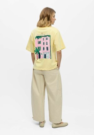 Femme aux cheveux mi-longs portant un t-shirt jaune avec un motif rose d’un bâtiment et d’un palmier, un pantalon beige à jambes larges et des chaussures plates beiges.