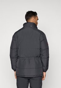 Casaco puffer preto com colarinho alto, punhos canelados e cintura com cinto. Apresenta um acolchoado horizontal e um pequeno logótipo na manga.