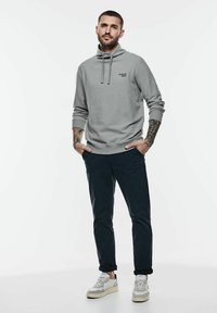 Světle šedá mikina s vysokým límcem, nastavitelnými šňůrkami a logem na přední straně. V kombinaci s tmavými kalhotami a bílými teniskami.