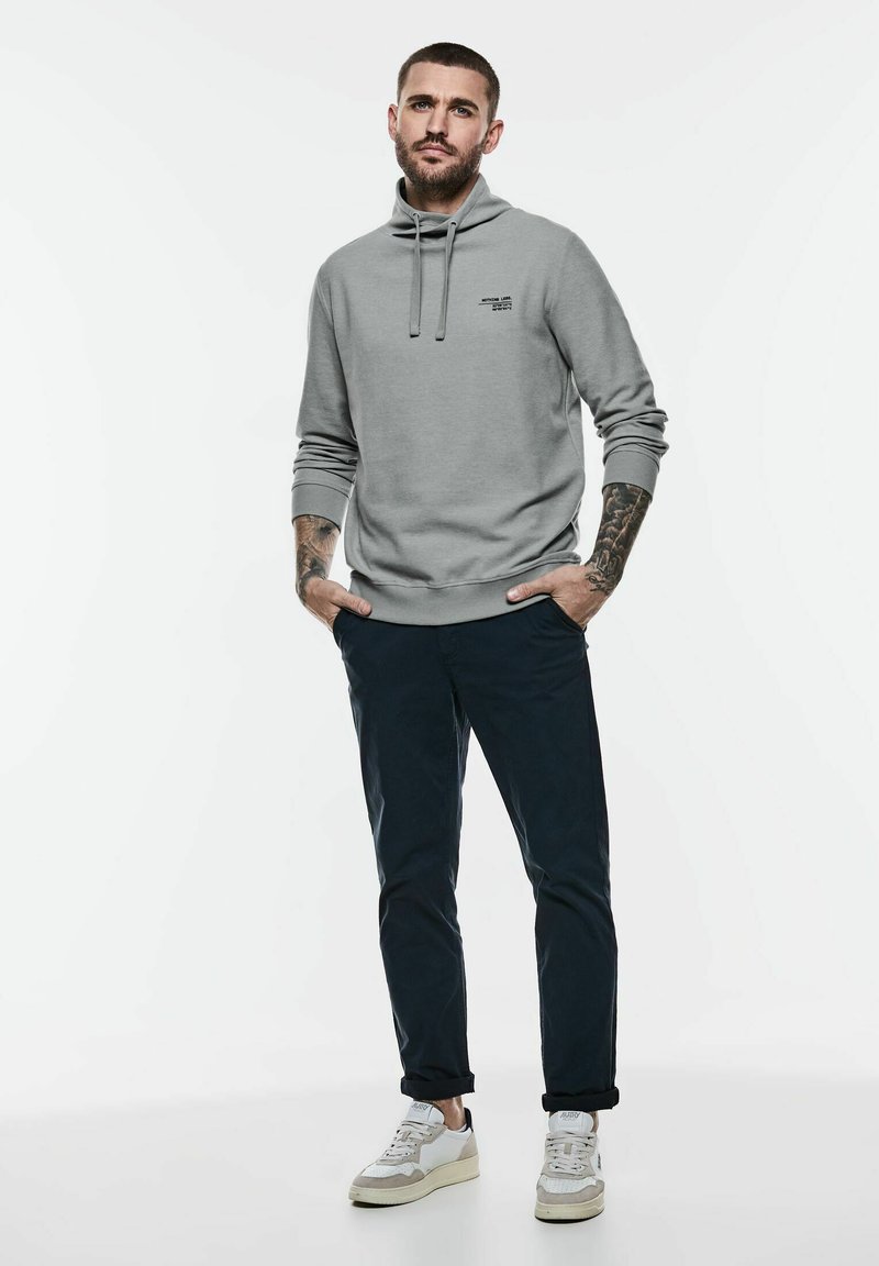 Světle šedá mikina s vysokým límcem, nastavitelnými šňůrkami a logem na přední straně. V kombinaci s tmavými kalhotami a bílými teniskami.