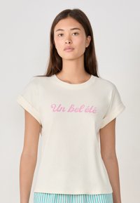 Camiseta de rayas en color crema con mangas enrolladas y texto rosa "Un bel été" impreso en el frente. La camiseta tiene un corte relajado y una textura suave.