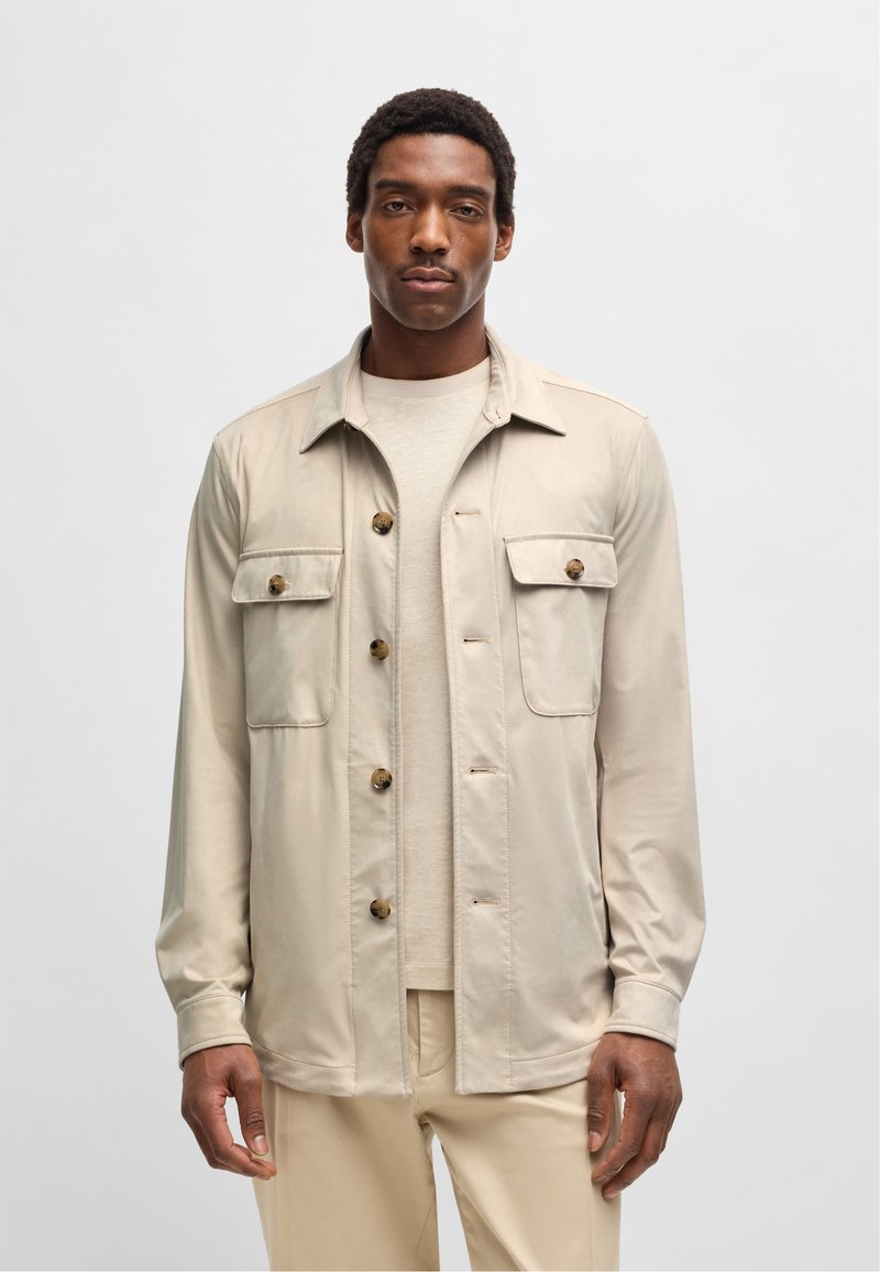 Lichtbeige button-up overhemdjasje met twee borstzakken, ronde kraag, lange mouwen en een gladde textuur. Gdragen over een lichtcrème top.