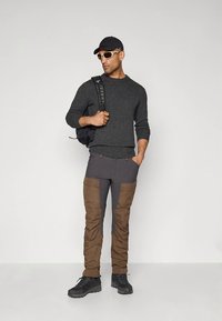 Dunkelgrauer Strickpullover, braune und graue Cargohosen mit Seitentaschen, schwarze Schuhe, schwarze Mütze und weiße Sonnenbrille. Legere Outdoor-Bekleidung.