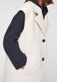 Cappa senza maniche di colore crema, con texture, colletto, due tasche e bottoni neri, indossata sopra una maglia a maniche lunghe scura.