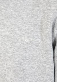 Sweatshirt gris en tissu doux, avec une légère texture et un logo brodé "SMILODOX" dans une teinte plus claire sur le devant.