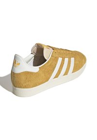 Hnědý semišový teniska s bílou gumovou podrážkou. Tři bílé pruhy na boku, polstrovaný jazyk a malé logo na patě.