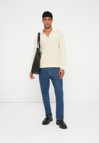 Maglione a coste beige chiaro con colletto zip, abbinato a jeans dritti blu scuro e mocassini neri, portato con una borsa a spalla nera.