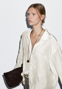 Light beige satin shirt with a loose fit, a deep V-neck, and buttons. Holding a dark brown suede clutch, complemented by a simple pendant necklace.