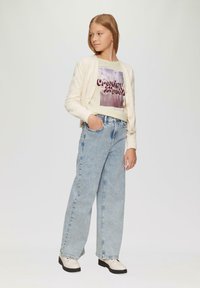 Helle Creme-Kardigan über einem grafischen T-Shirt mit lila Text, kombiniert mit hochgeschnittenen, hellblauen Jeans und cremefarbenen Schuhen mit schwarzen Sohlen.