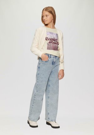 Jasna kremowa kardigan nałożona na t-shirt z grafiką z fioletowym tekstem, w połączeniu z jasnoniebieskimi jeansami o wysokim stanie i kremowymi butami z czarnymi podeszwami.