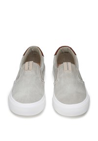 Scarpe slip-on in tela grigio chiaro con superficie texturizzata e suola in gomma bianca. Presentano un accento marrone sul tallone e un'etichetta sulla linguetta.