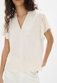 Blusa leggera color crema con scollo a V, maniche corte a volant e un leggero motivo testurizzato, abbinata a pantaloni chiari.