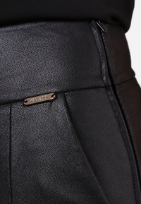 Pantalons en simili cuir noir avec une texture lisse, présentant une plaque logo dorée sur la ceinture et des détails subtils sur les coutures.