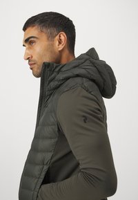 Olijfgroene gewatteerde vest met een ritssluiting aan de voorkant en een capuchon. Beschikt over een soepele textuur en een logodetail op de bovenarm.