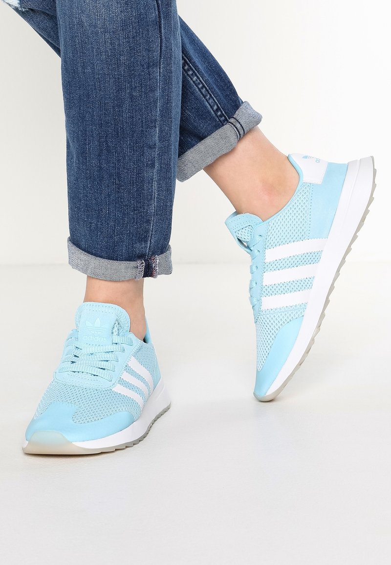 Light blue sneakers with mesh and synthetic details, featuring three white stripes, a rounded toe, and a textured sole. Denim cuffed pants.