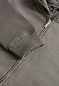 Pelēka zip-aizdare hoodei, kas izgatavota no mīkstas auduma. Iezīmes riboti aproces un metāla rāvējslēdzēja vilcējs. Iekšējā birka norāda, ka izmērs ir vidējs.