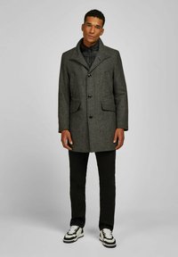 Grau-karierter Überwurf mit Kragen, zwei vorderen Taschen und vier Knöpfen, getragen über einer schwarzen Daunenjacke und kombiniert mit schwarzen Hosen und Sneakers.