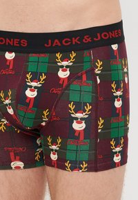 Boxers de Noël décorés en bordeaux avec un motif de rennes, des accents en tartan et le texte "Noël". Ils possèdent une ceinture élastique noire.