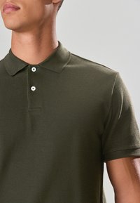 Maslinasto zelena polo majica od teksturiranog tkanja; ima klasični ovratnik, dvije gumbice na prednjoj strani i kratke rukave. Suptilan rebrasti uzorak.