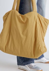 Borsa tote in tessuto giallo con una texture rugosa, manici larghi e una forma morbida, indossata con jeans blu e scarpe da ginnastica argentate.