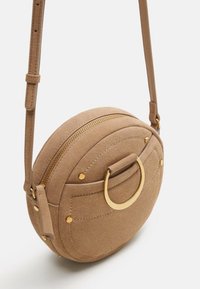 Okrąła, beżowa torebka typu crossbody z zamszu, z akcentem w postaci złotego metalowego pierścienia, zamknieniem na zamek oraz przeszyciami. W zestawie regulowany pasek.