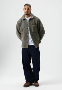 Fleece jacket in olive green with a textured finish, featuring a collar and two front pockets. Paired with loose-fitting dark denim pants.