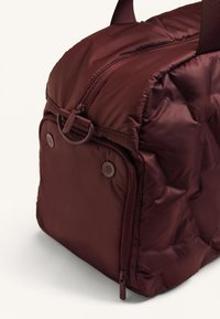 Burgundi hűtőtáska cipzárral, kerek formájú, texturált anyagból készült. Két oldalsó gombbal és egy hurokkal a csatlakoztatáshoz rendelkezik.