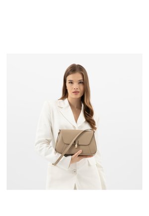 Vrouw in wit colbert die een beige lederen handtas met schouderband vasthoudt tegen een witte achtergrond.