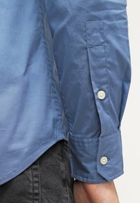 Gros plan sur un poignet de chemise bleu à manches longues avec deux boutons blancs, porté avec un pantalon gris foncé.