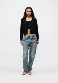 Cardigã curto de malha preta com fecho de botão sobre um top cropped correspondente, combinado com jeans claros rasgados e sapatilhas bege.