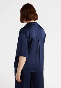 Maglietta a maniche corte blu navy in tessuto satinato liscio. Presenta un colletto, una vestibilità ampia e una piega sul retro per una maggiore texture e movimento.