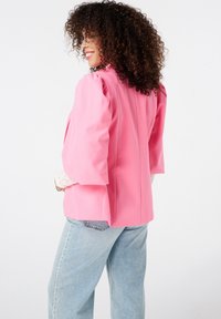 Roze blazer met opbollende mouwen, gestructureerd ontwerp en een getailleerde pasvorm. Gecombineerd met lichtgewassen denimjeans. Soepele stof zonder zichtbare patronen.