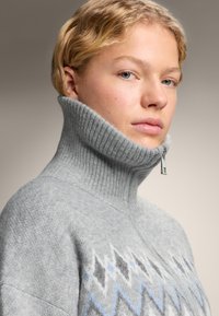 Šedý svetr s vysokým límcem z měkkého pleteného materiálu, zdobený abstraktním světle modro-šedým geometrickým vzorem a zipem u límce.