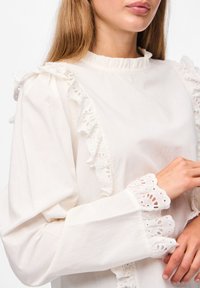 Blusa blanca con bordes de encaje bordado en los hombros y puños. Presenta un escote con volantes y mangas largas en una tela de textura suave.