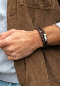 Bracelet en cuir tressé marron avec un fermoir métallique sur un poignet, associé à une veste en velours côtelé marron texturée et une chemise grise.