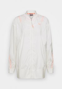 Valkoinen, kevyt harjoitusjakku, jossa on täyspitkä vetoketju, ribatut hihat ja neonoranssit yksityiskohdat hartioilla. Sujuva pinta.