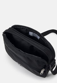 Černá polyesterová crossbody taška s dvojitými zipovými přihrádky a značkovou visačkou uvnitř. Obsahuje nastavitelný popruh pro pohodlné nošení.