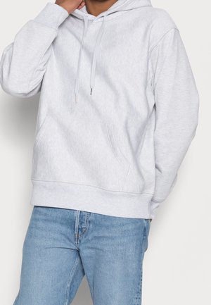 Persoon die een lichtgrijze hoodie met voorvak draagt en een blauwe spijkerbroek, met één hand aan het gezicht.