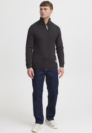 Pull en tricot noir avec un col montant, comportant une fermeture éclair quart et des poignets côtelés. Associé à un jean bleu et des baskets de couleur claire.