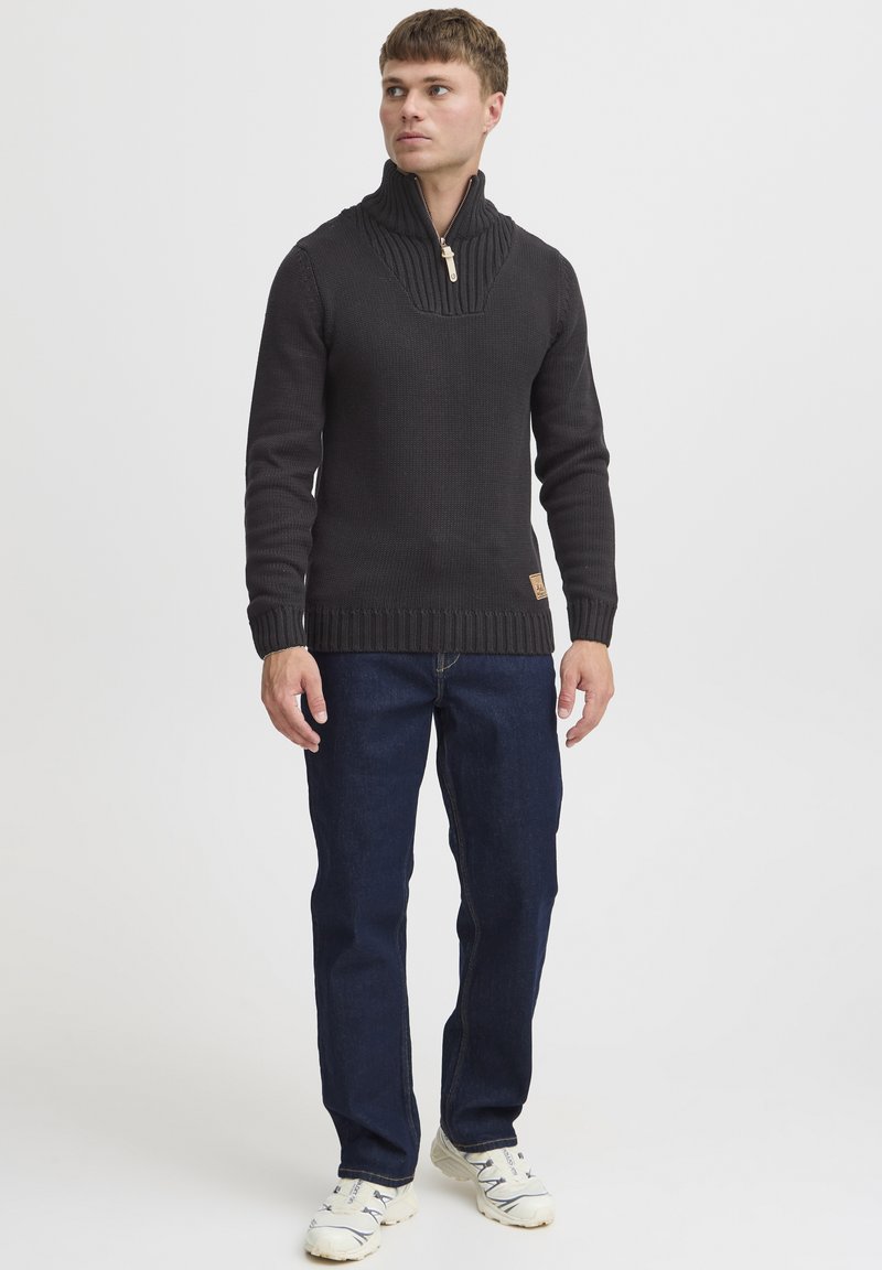 Dunkel gestrickter Pullover mit hohem Kragen, versehen mit einem Viertel-Reißverschluss und gerippten Bündchen. Kombiniert mit blauen Jeans und hellen Sneakers.