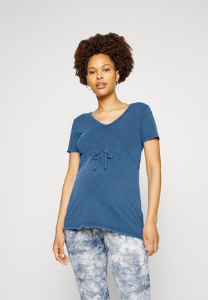 Blusa de maternidade em mistura de algodão azul, com decote em V, mangas curtas e um detalhe de laço na frente. Combinada com leggings estampadas em azul e branco.