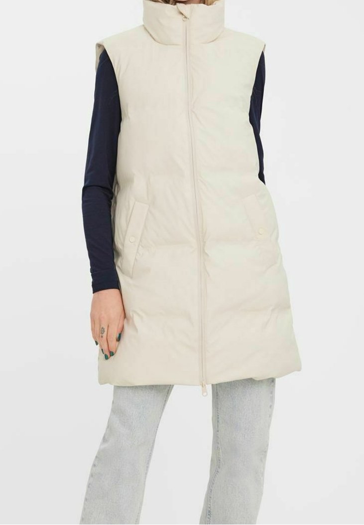 Veste sans manches beige matelassée avec col montant, fermeture éclair à l'avant et deux poches à bouton-pression, portée par-dessus une chemise à manches longues foncée.