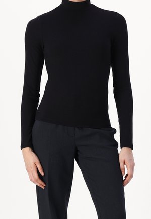 Frau trägt ein enganliegendes schwarzes Langarm-Longsleeve mit Rollkragen und dunkle Hose, steht mit entspannten Händen an den Seiten.