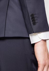 Navy colbert met een mouw met vier zwarte knopen en een subtiele textuur, gecombineerd met een witte bloesmanchet met afgeronde rand en knoopsluiting.