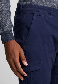 Marineblauwe cargobroek met een zijzak met knoopsluiting en gestructureerde katoenen stof, gecombineerd met een grijze gebreide trui mouw.