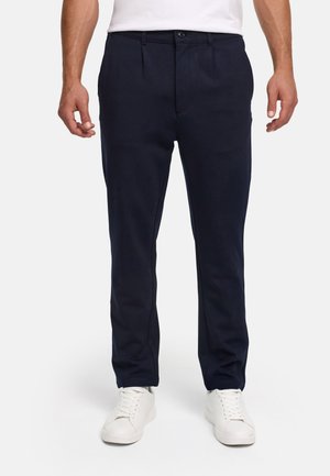 Uomo con pantaloni neri slim fit, sneaker bianche e camicia bianca infilata, in piedi su uno sfondo chiaro uniforme.