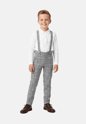Dziecko ubrane w białą koszulkę polo z długim rękawem, szare szelki w kratę, szare spodnie w kratę oraz brązowe buty, stojące w neutralnej pozycji.